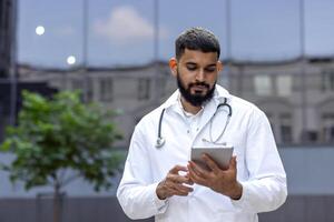 A young Muslim male doctor stands near the clinic and uses a tablet, chats with a patient, writes down the history of illness and treatment. photo