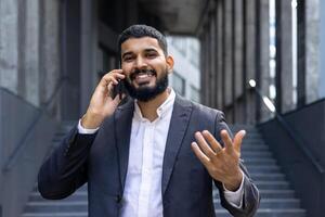 retrato de un contento joven indio empresario hablando en el teléfono en el calle, gesticulando con su manos y sonriente a el cámara. foto