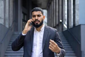 Upset young Indian male businessman talking on the phone on the street near the office, courtroom. Dissatisfied and gesticulating with his hands. photo