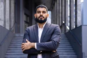 Portrait of a young Muslim male lawyer standing in a business suit outside a courthouse and looking confidently into the camera with his arms crossed. photo
