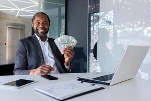 de cerca retrato de un afroamericano empresario sentado en el oficina a un escritorio, participación un ventilador de dinero en efectivo, señalando y sonriente a el cámara. foto