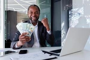 Happy African American businessman sitting in modern office holding stack of dollars in hands, salary increase, winning money in lottery, looking at camera. photo