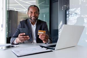 mayor africano americano hombre en traje sentado a escritorio en oficina. él es participación un crédito tarjeta y un teléfono. exitosamente pagado en línea compras, pedidos, facturas. contento mirando a cámara. foto