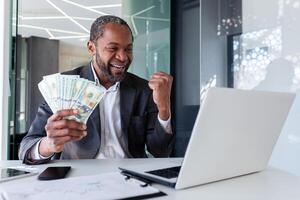 Happy african american man sitting in office at table in front of laptop, holding money cash in hands and enjoying success showing victory gesture with hand. photo