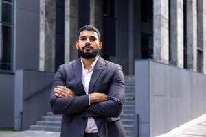 Close-up portrait of a young Muslim, male lawyer in a business suit standing seriously outside a courthouse and looking confidently into the camera, with his arms folded. photo