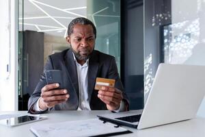 mayor enojado africano americano hombre en traje sentado a escritorio en oficina. él es participación un crédito tarjeta y un teléfono. sin éxito pagado en línea compras, pedidos, facturas. triste mirando a el cámara. foto