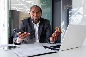 Portrait of a worried African American businessman sitting in the office at a desk, holding a credit card and a phone and looking disappointedly at the camera and throwing up his hands. photo