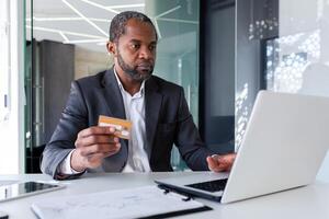 Serious thinking man in business suit working with laptop inside office, boss holding bank credit card, online purchase and money transfer. photo