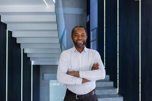 Mature african american businessman with crossed arms smiling and looking at camera inside office building, satisfied and successful man at work in shirt. photo