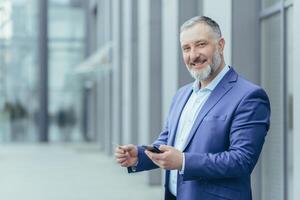 Portrait of successful gray-haired businessman man smiling and looking at camera, boss holding bank credit card and smartphone, director using money transfer app photo