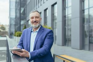 Portrait of successful senior gray-haired businessman boss, man outside office using tablet computer smiling and looking at camera, investor in business suit, happy banker photo