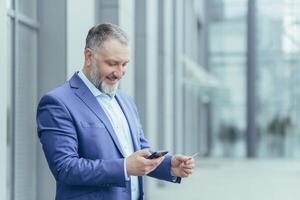 Senior gray-haired businessman outside office building, man in business suit uses mobile phone smartphone to buy in online store and order service, holds bank card in hands photo