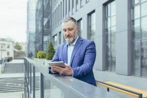 Successful and serious pensive senior gray haired man outside office building using tablet computer, businessman investor reading news from tablet photo