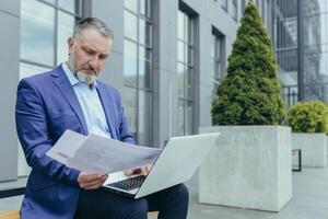 grave mayor canoso empresario inversor con papeleo sentado en banco fuera de oficina edificio, hombre en negocio traje con documentos y ordenador portátil trabajando foto
