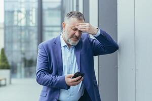 Senior gray-haired businessman sick outside office building, man holding hand to head, having severe headache, boss trying to call doctor on phone and call ambulance photo