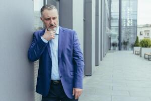 Senior gray haired man outside office building, standing and thinking, confused and sad businessman boss in business suit, over decision photo