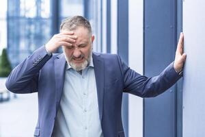 Senior gray haired man businessman upset and desperate outside office building, holding head with hands, mature boss bankrupt photo