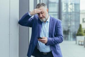 Senior gray-haired businessman in despair upset reading bad news on phone, man outside office building in business suit, bankrupt investor lost money, banker in depression photo