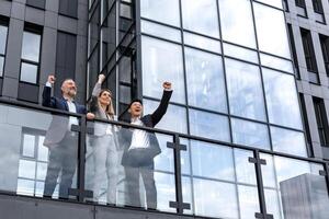 Diverse business group of people colleagues in business clothes outside office building, male and female employees rejoice and celebrate victory hold hand up and smile successful team photo