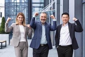 Successful diverse business team, man and woman outside office building smiling and looking at camera, portrait of business people in business clothes, colleagues holding hands up and rejoicing, victory gesture photo