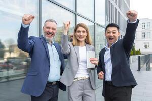Successful diverse business team, business people looking at camera and rejoicing in success and victory, man and woman holding hands up outside office photo