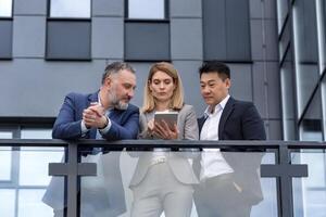 Three colleagues, diverse business group, business people outside office building talking and discussing work process, looking at tablet computer thoughtfully photo