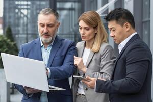 Diverse business group of three people talking and discussing strategy outside office building businessman and businesswoman looking at laptop screen while standing photo