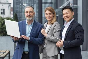 Portrait of three diverse business colleagues, business group outside office building, asian man and woman looking at camera and smiling, dream team with laptop photo