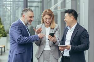 Diverse business team, male and female business people together outside office, group of successful business people looking at colleague's phone, smiling and happy photo