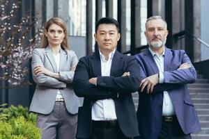 Asian boss with team, portrait of group of people with crossed arms looking at camera, diverse business people outside office building standing focused photo