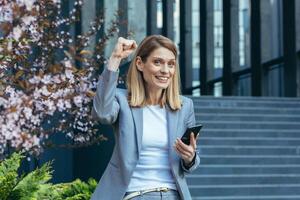 Successful business woman in a business suit outside the office reads good news from the phone, smiles and rejoices in victory, a gesture of triumph and victory photo