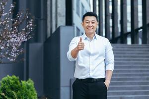 Happy asian agent businessman looking at camera and smiling holding thumbs up outside office photo