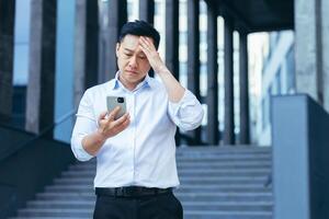 Upset Asian businessman reads news from phone screen, man disappointed with bad news photo