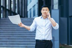 Frustrated insurance agent with documents talking on the phone outside the office, depressed Asian businessman photo