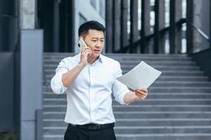 Frustrated insurance agent with documents talking on the phone outside the office, depressed Asian businessman photo