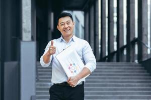 Portrait of Asian businessman broker, man outside office holding laptop and papers with charts, looking at camera smiling holding thumbs up photo