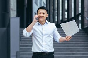 Happy and successful Asian businessman talking on the phone, holding documents outside the office photo