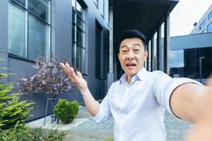 Asian talking on call uses phone, looks at camera and cheerfully waves hands greeting gesture photo