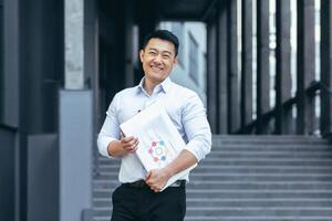 Portrait of happy asian business man, accountant with financial report and laptop, looking at camera and smiling outside office photo