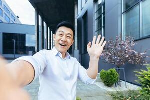 Asian talking on call uses phone, looks at camera and cheerfully waves hands greeting gesture photo