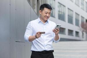 Asian businessman smiling and happy doing online shopping outside office building, man using mobile phone, holding bank credit card photo