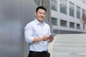 Successful asian businessman with tablet computer in hands, smiling and looking at camera, man standing outside office building photo