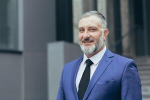 Close up photo portrait of successful and senior experienced businessman, boss smiling and looking at camera, man in business suit standing outside office building