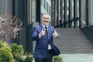 Successful senior gray haired businessman boss, outside office building smiling and looking at camera, experienced investor showing middle finger up, man in business suit photo