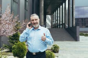 Senior gray-haired businessman in a shirt outside an office building talking on the phone, walking around the city photo