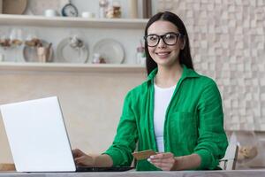 Online shopping. Portrait of a beautiful woman in glasses and a green shirt sits at home with a laptop, holds a credit card in her hands. Makes purchases on the Internet, orders delivery, pays online. photo