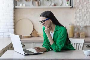 Online shopping. A young beautiful woman in glasses and a green shirt sits at home with a laptop, holds a credit card in her hands. Makes purchases on the Internet, orders delivery, pays online. photo