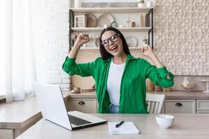 Portrait of happy young business woman in glasses and green jacket. She sits at the table at home, works with a laptop, is happy, raised her hands up, received good news, smiles. photo