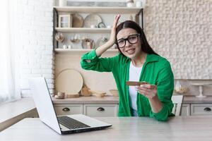 Monetary crisis. Credit debt. Upset young woman in glasses and green shirt holding credit card, sitting at laptop at home, holding head, shocked, confused. photo