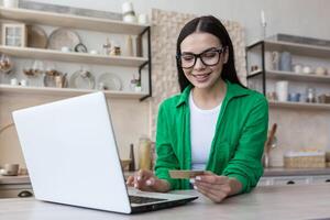 Online shopping. A young beautiful woman in glasses and a green shirt sits at home with a laptop, holds a credit card in her hands. Makes purchases on the Internet, orders delivery, pays online. photo